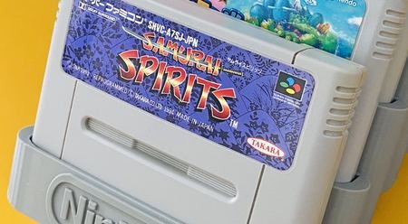 Gros plan d'une cartouche Samurai Spirits maintenue par les flancs dans son encoche